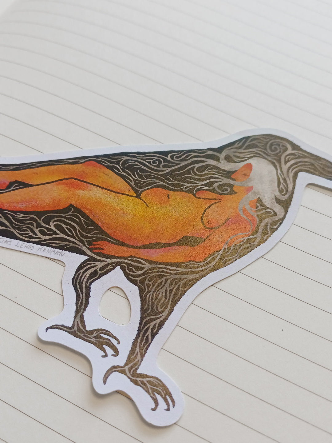 A die-cut sticker featuring a stylized raven with a nude human figure inside, printed in orange, brown, and black, shown on a cream-colored lined notebook page.