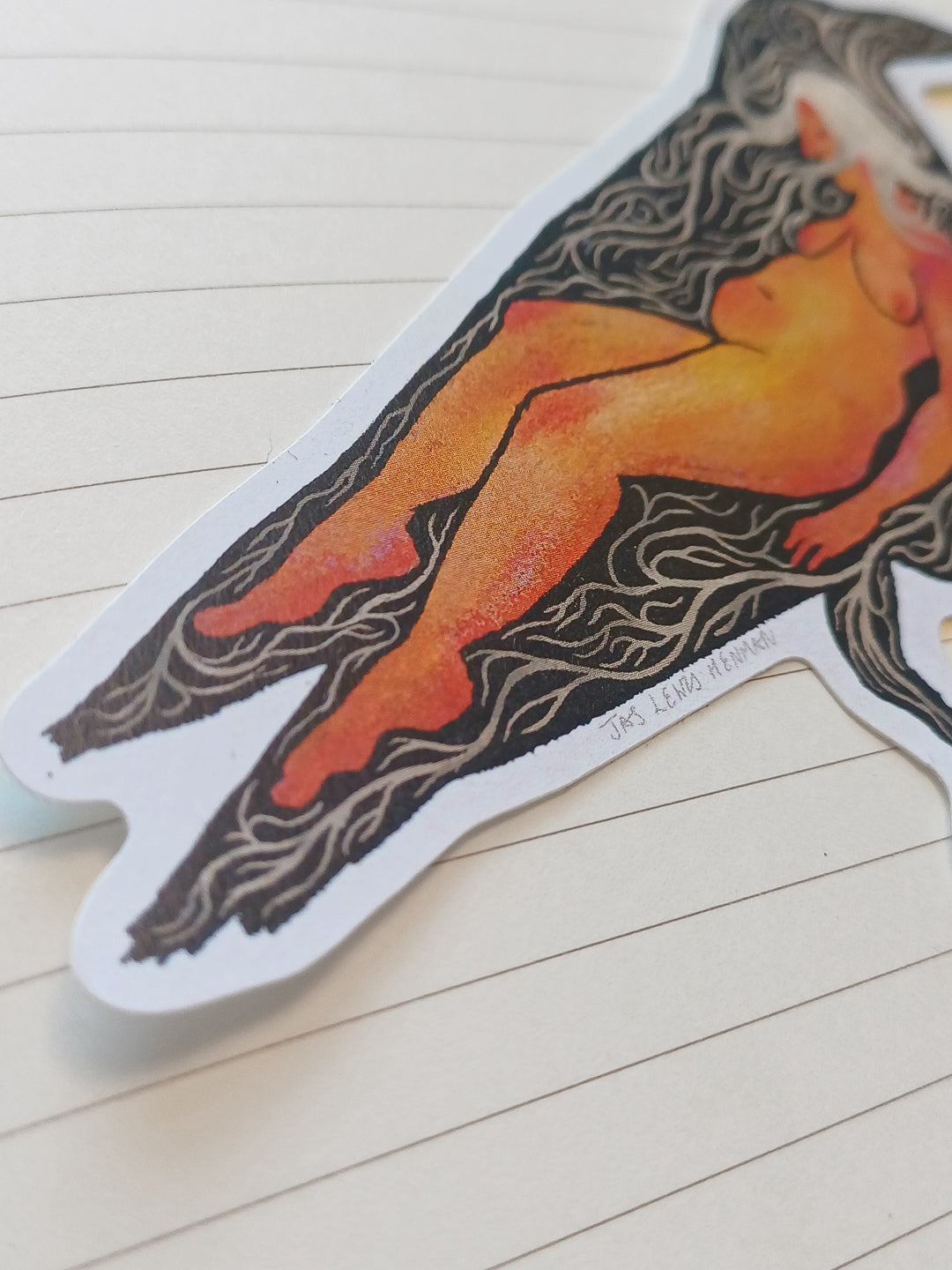 A die-cut sticker featuring a stylized illustration of a nude female figure in orange and yellow tones with black hair, placed on a cream-colored lined notebook page.