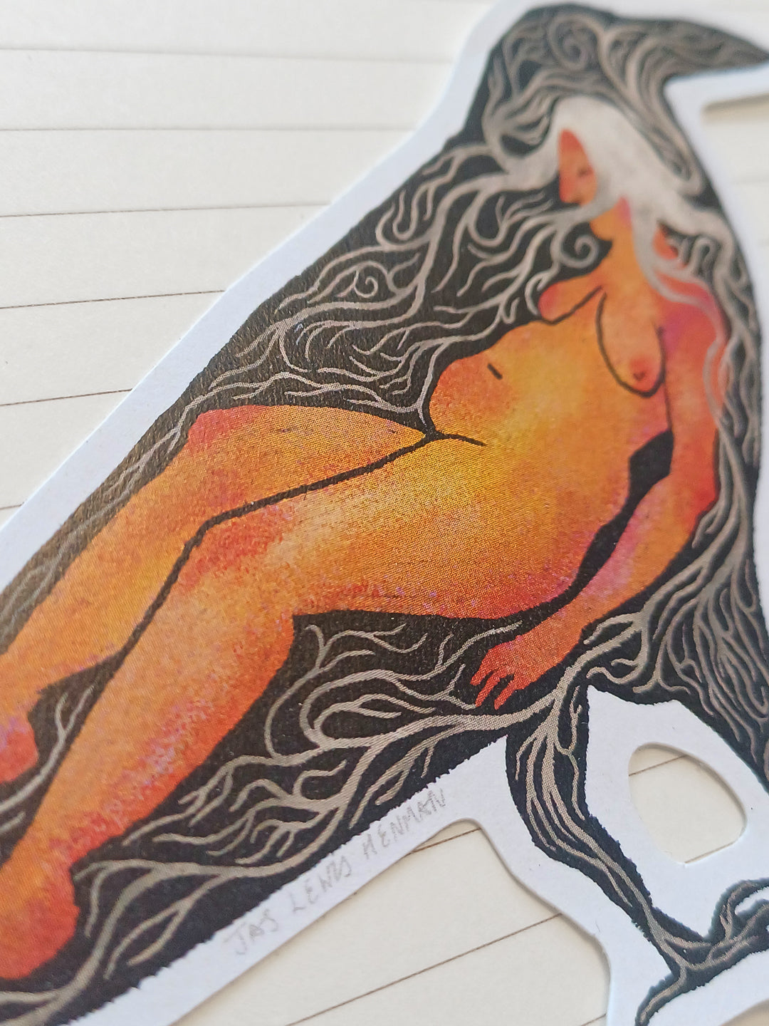 A die-cut sticker featuring a stylized illustration of a nude female figure with flowing dark hair, printed on a white backing and photographed on a light wood surface.