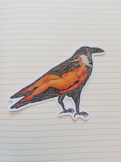 A die-cut sticker of a raven with a nude female figure inside, detailed with black line art and filled with orange, red, and yellow tones, placed on a cream-colored lined notebook page.
