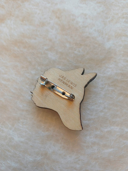 A wooden pin shaped like a fox head, showing the back side with a metal safety pin c-clasp and engraved text reading 'JAS LEWIS HENMAN', placed on a soft, textured cream-colored fabric background.