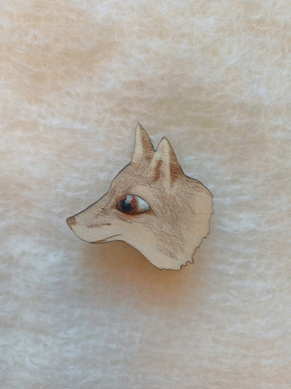 A small wooden fox head pin badge brooch with a detailed engraved face and brown woollen needle felted eye, shown on a soft beige textured fabric background.
