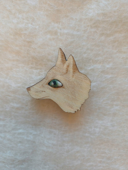 A small wooden fox head brooch with a detailed green wool eye, shown on a textured beige fabric background.