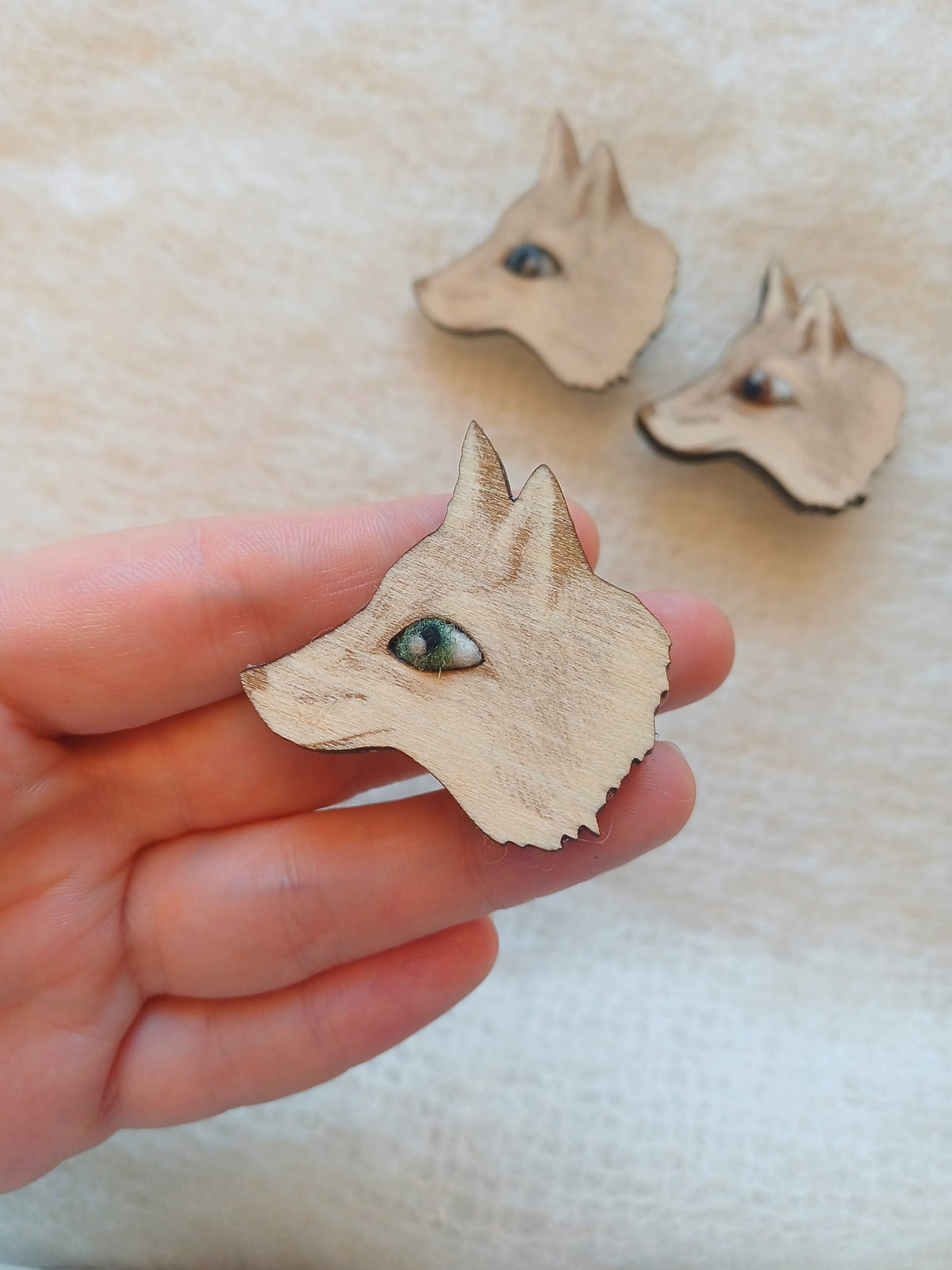 A hand holds a small wooden fox head brooch with a green eye, with two similar wooden fox head accessories blurred in the background on a light beige textured surface.