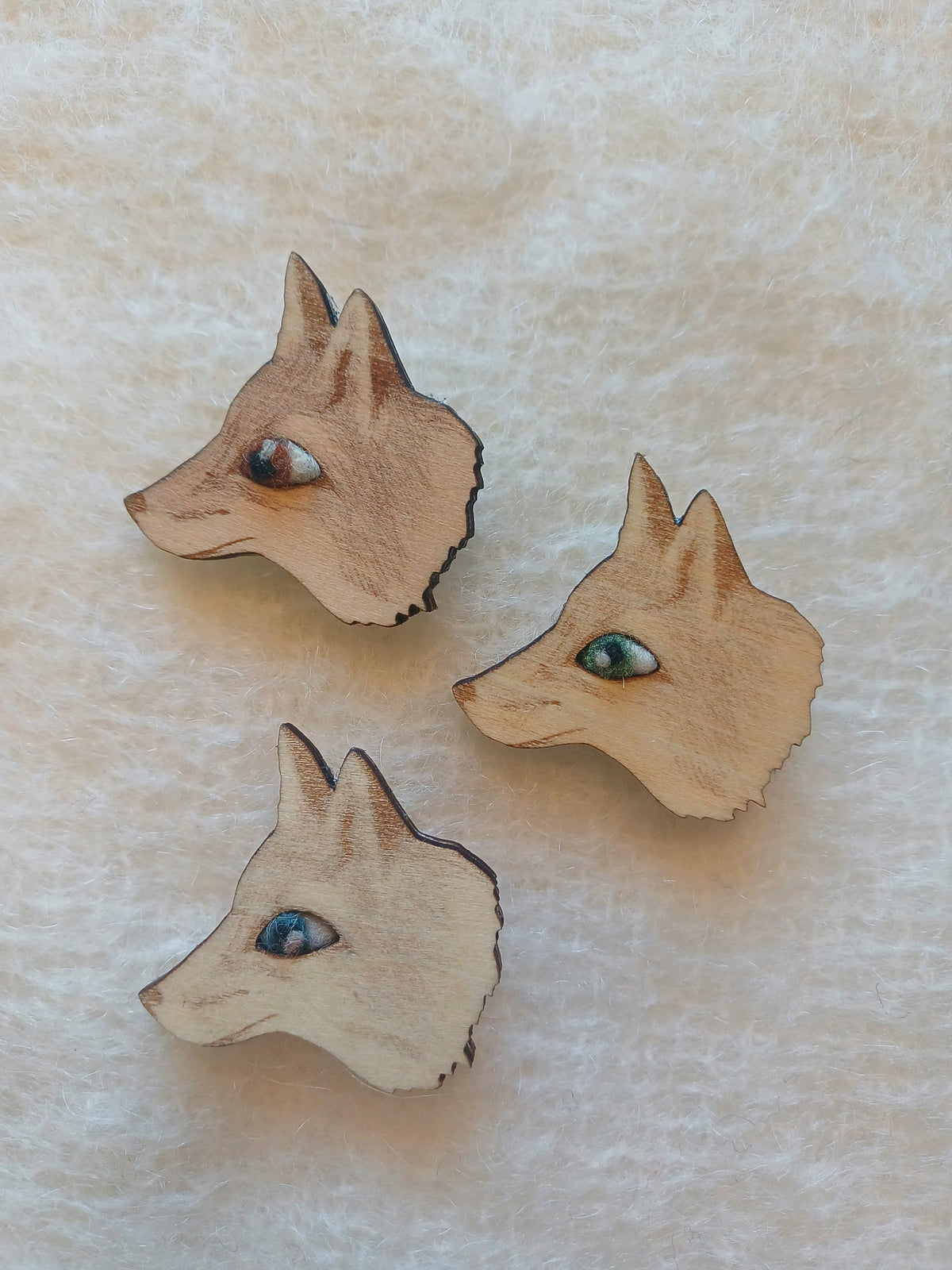 Three small wooden laser cut fox head brooches with woll needle felted eyes in brown, green, and blue, arranged on a textured off-white surface.