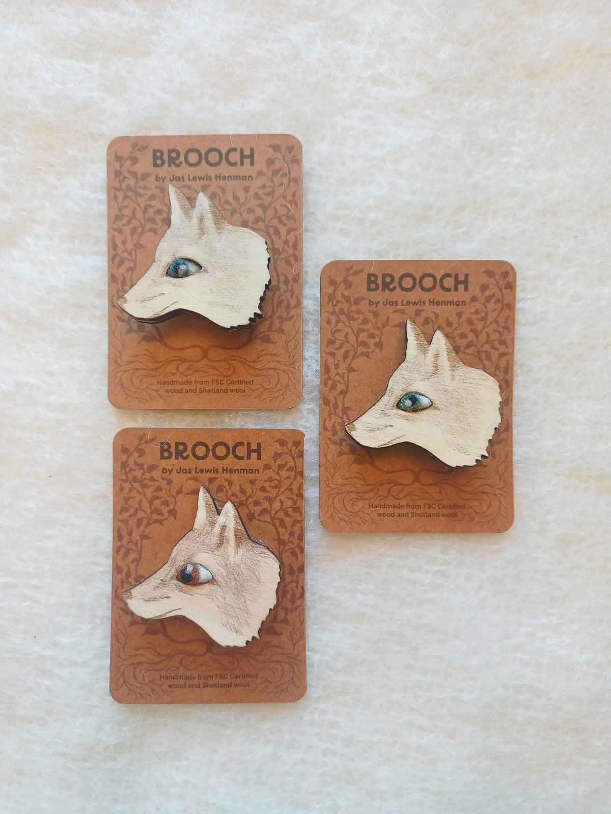 Three small brooches shaped like fox heads are displayed on brown backing cards on a light textured fabric background. The cards read BROOCH and show the brand as Jas Lewis Henman
