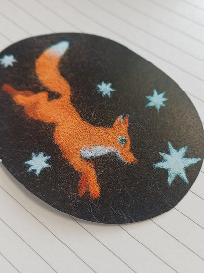 An oval sticker featuring an orange felt fox with white star accents on a black background, decorated with light blue stars, shown on a white lined notebook page.