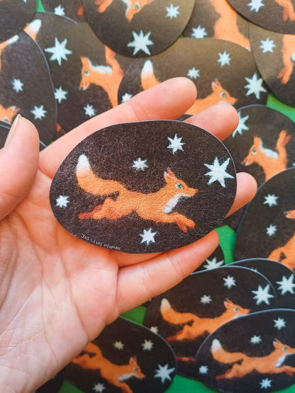 A hand holds a dark brown oval sticker featuring an orange fox running across a black background with white star details. Multiple identical patches are scattered in the background on a green surface.