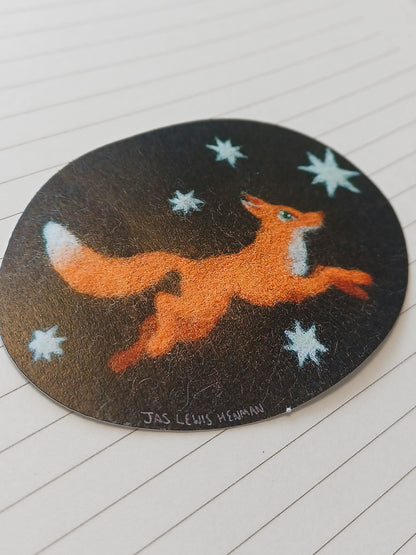 A circular black sticker featuring a felt orange fox running across a field of white stars, placed on a white paper surface.