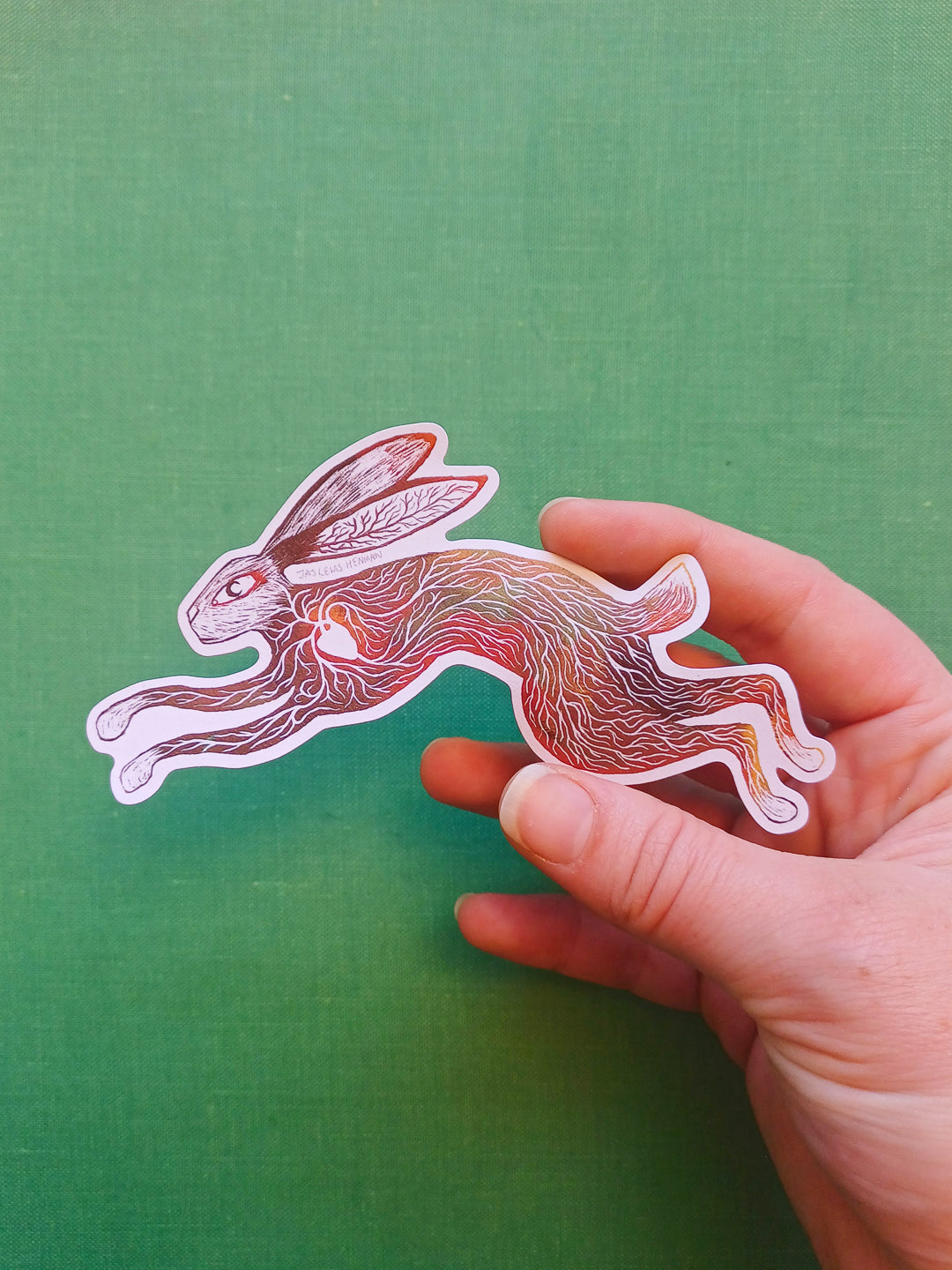 A hand holding a die-cut sticker of a leaping rabbit with intricate red, orange, and brown line art on a white border, set against a textured green background.