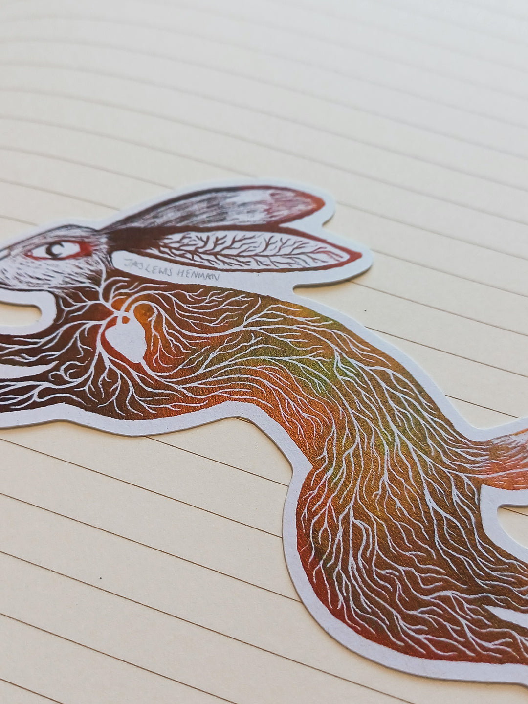 A die-cut sticker of a rabbit with intricate white line-art patterns and a warm gradient fill of orange, brown, and green, placed on a beige lined notebook page.