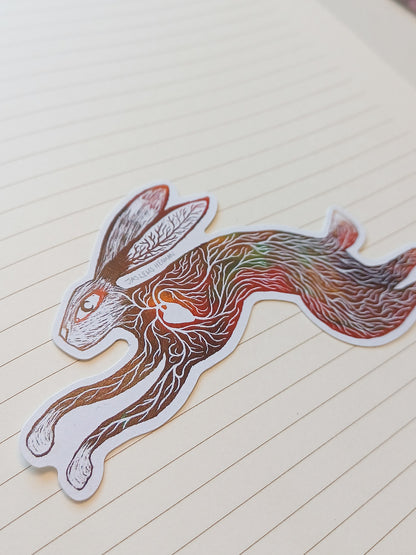 A die-cut sticker of a leaping rabbit filled with intricate brown, red, and green line art patterns, placed on a cream-colored lined notebook page.