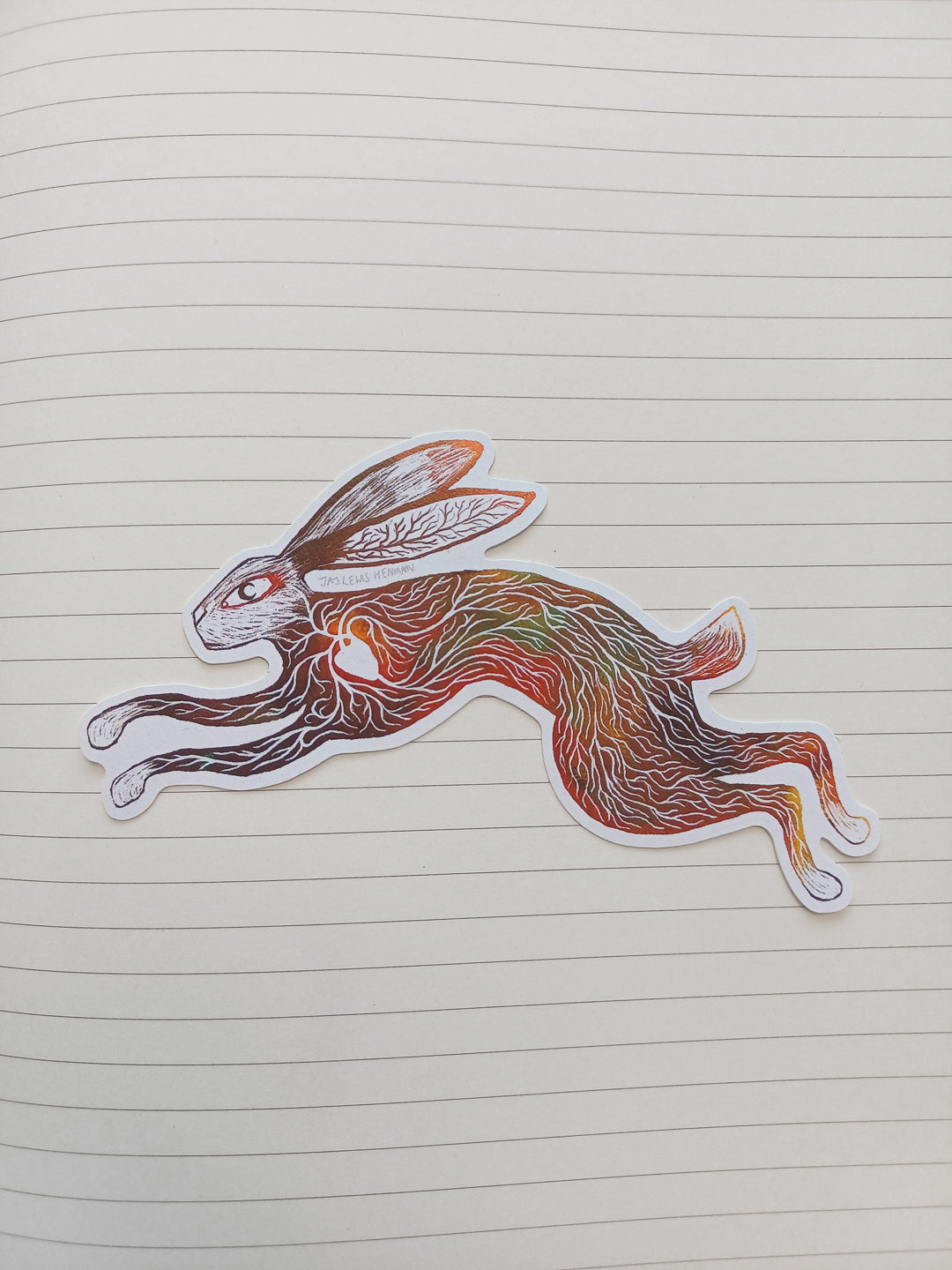 A die-cut sticker of a leaping rabbit with intricate multicolored line art, placed on a sheet of lined notebook paper.