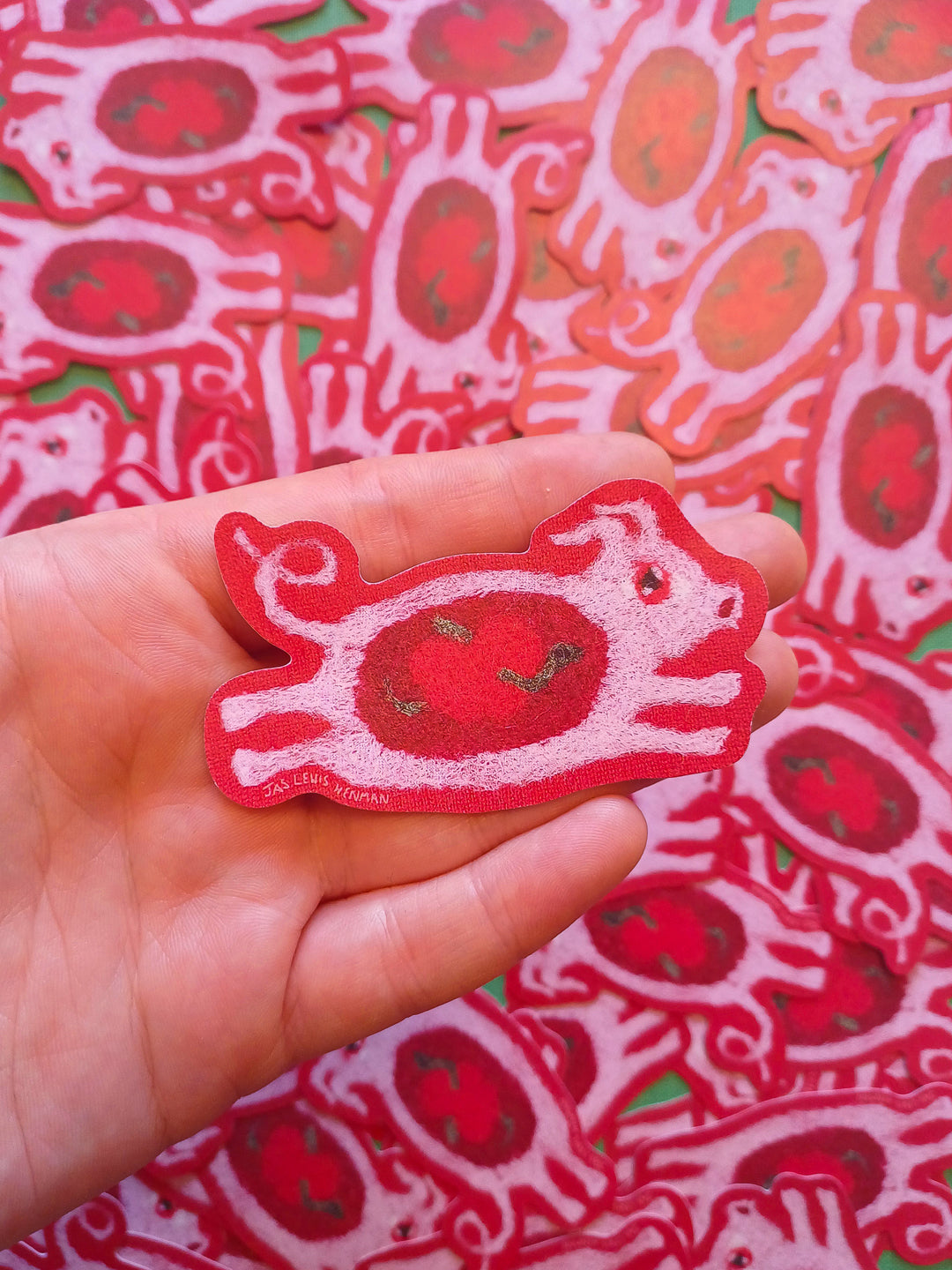 A hand holds a red and pink sticker of a  felt leaping pig, with a background of many similar stickers piled together.