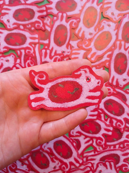 A hand holds a red and pink sticker of a  felt leaping pig, with a background of many similar stickers piled together.