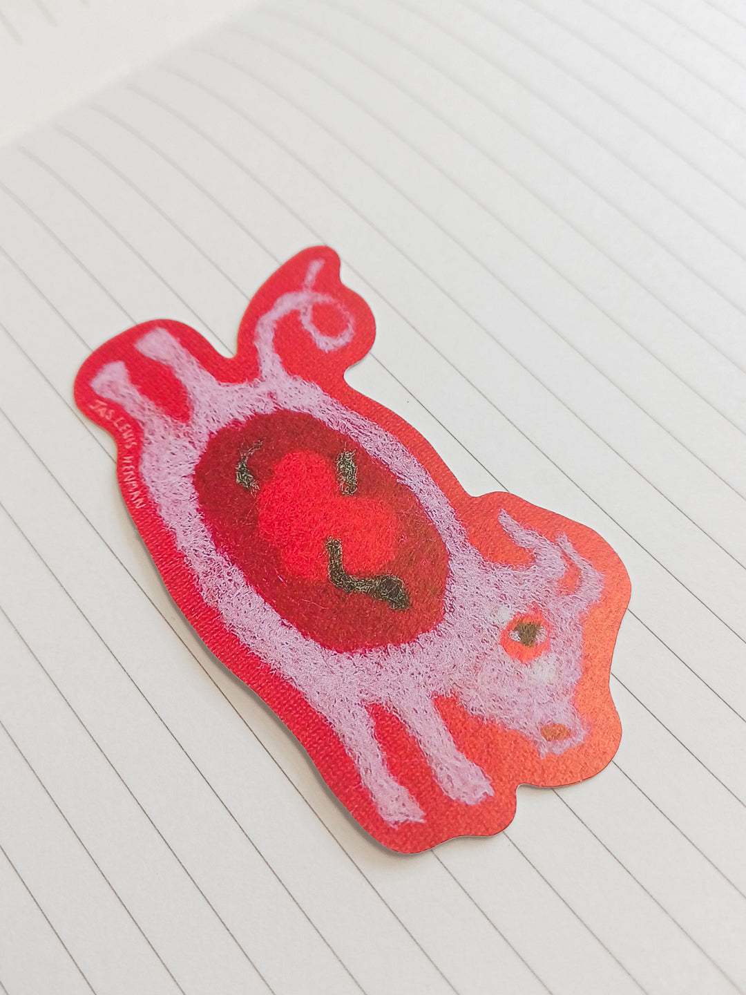 A red and pink textured sticker shaped like a stylized pig, placed on a white lined notebook page.