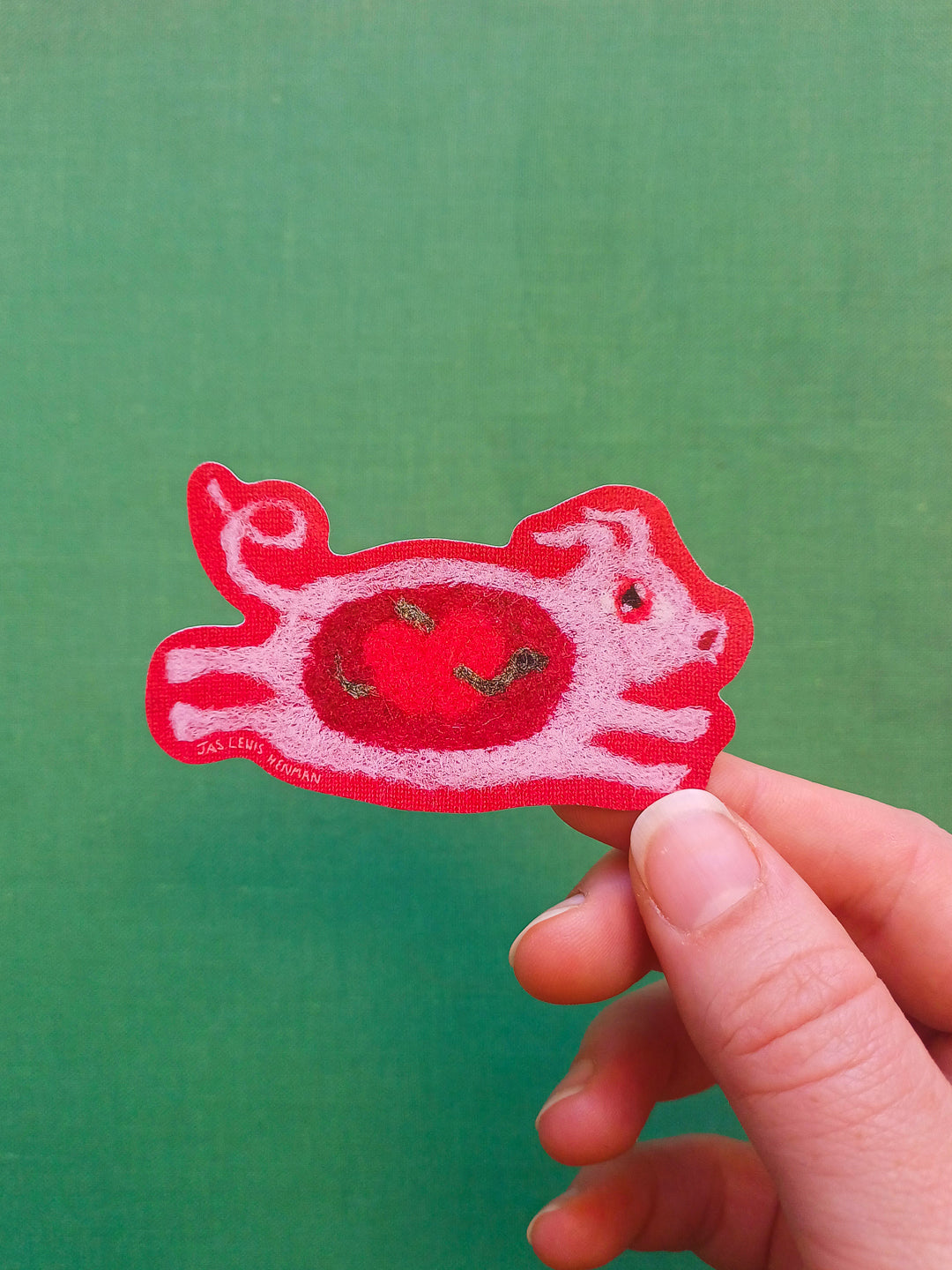 A hand holding a red and pink sticker from a felt design shaped like a leaping pig with a red apple in it's belly, set against a solid green background.