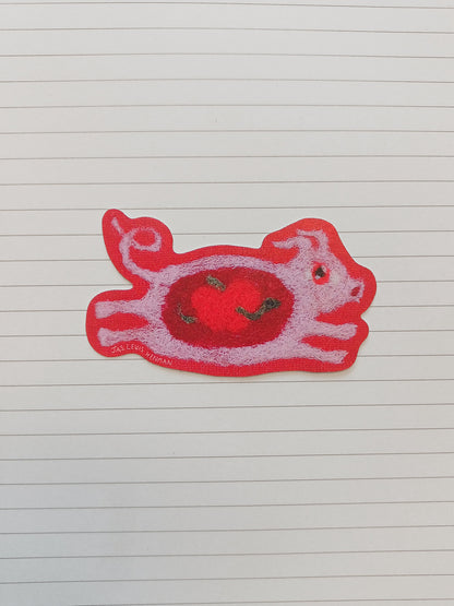 A red and pink sticker shaped like a stylized pig with an apple on its chest, placed on a white lined notebook page.