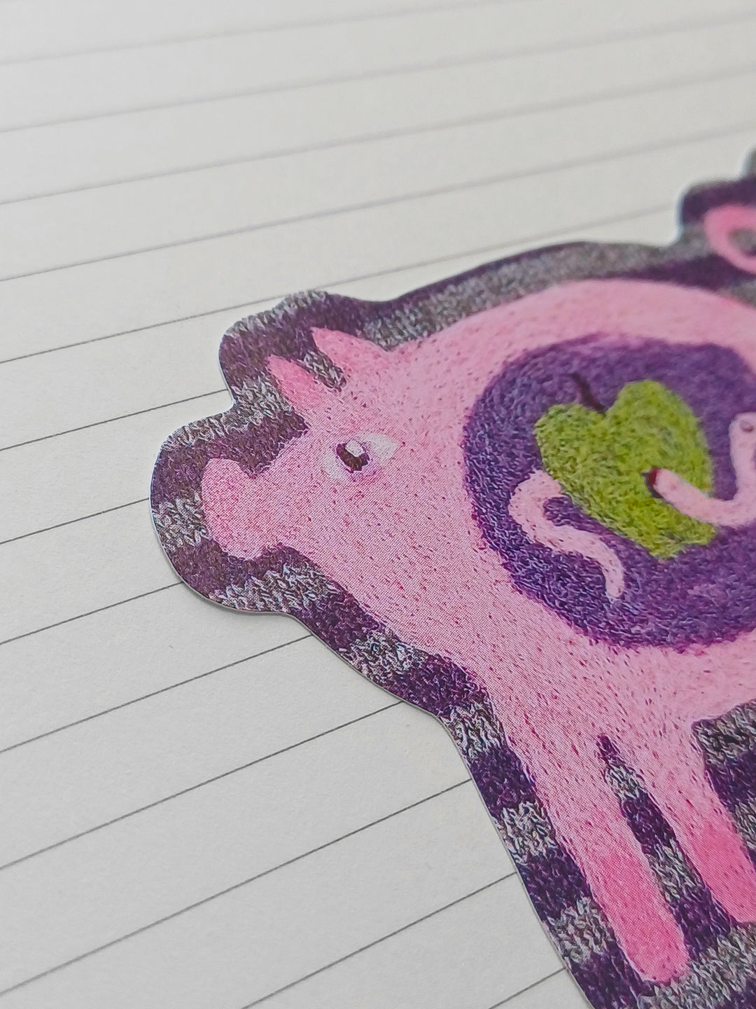 A close-up of a pink needle felted pig sticker with a purple and gray striped border and a green apple detail, placed on a white lined paper background.