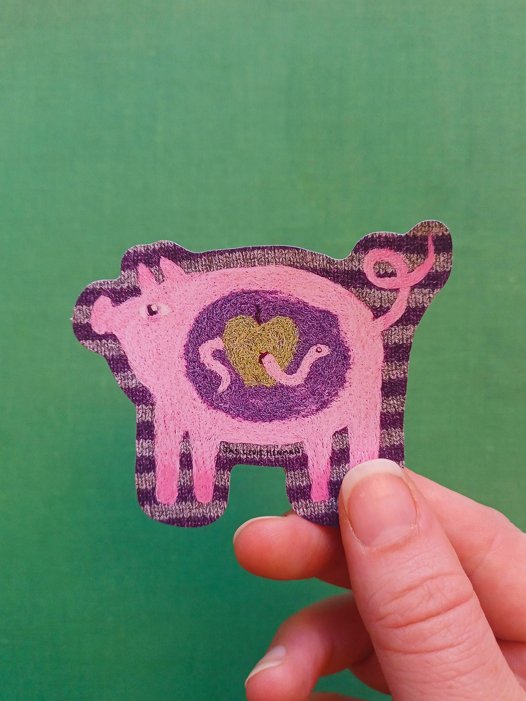 A hand holding a pink and purple pig-shaped patch or sticker with a green textured center, set against a solid green background.
