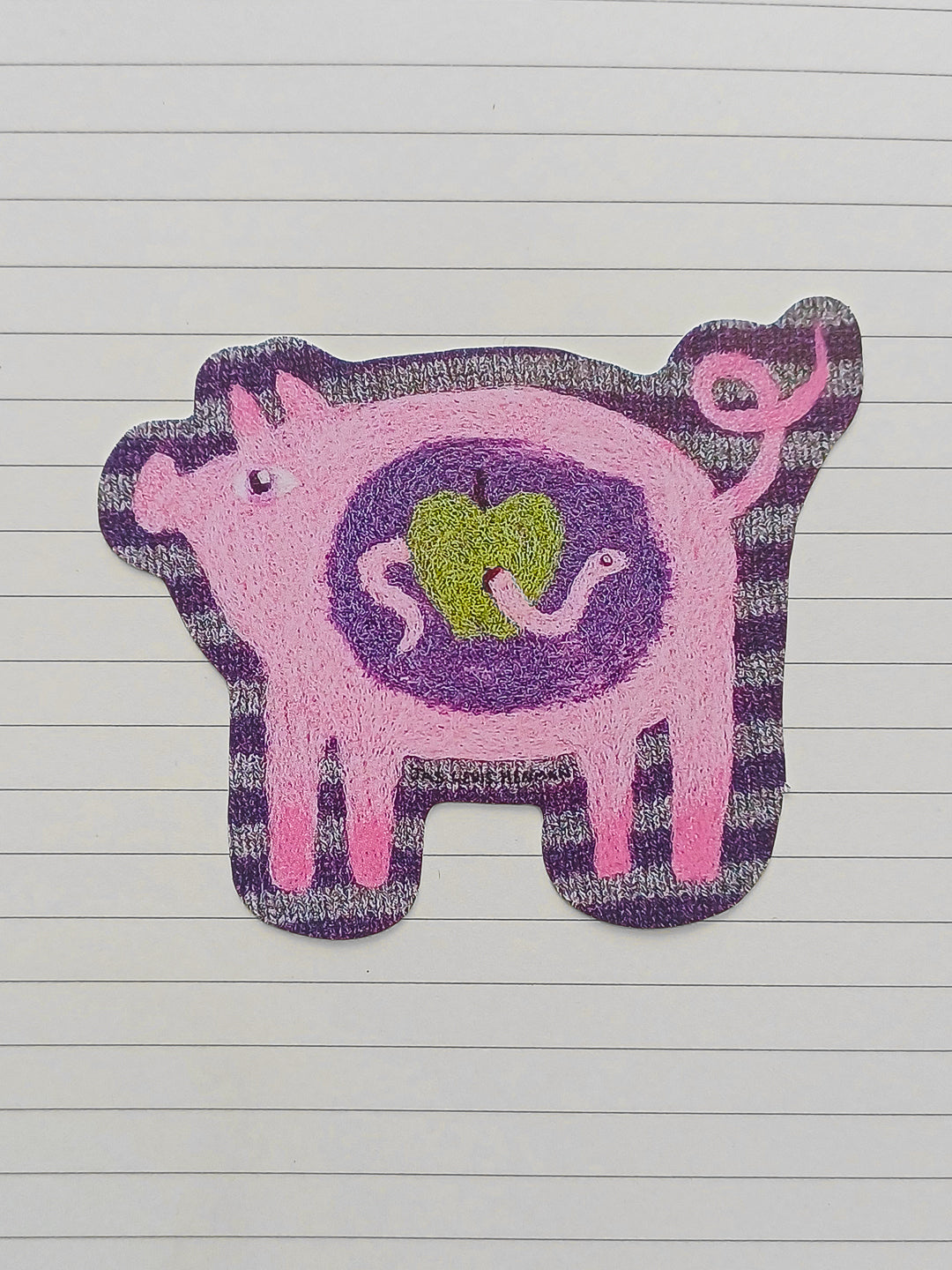 A pink and purple pig-shaped sticker with a green apple design in the center, placed on a white lined notebook page.