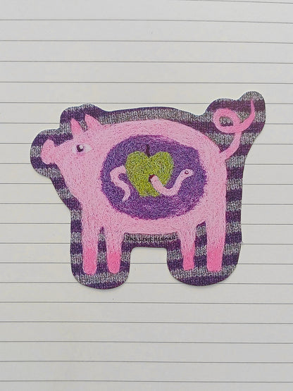 A pink and purple pig-shaped sticker with a green apple design in the center, placed on a white lined notebook page.