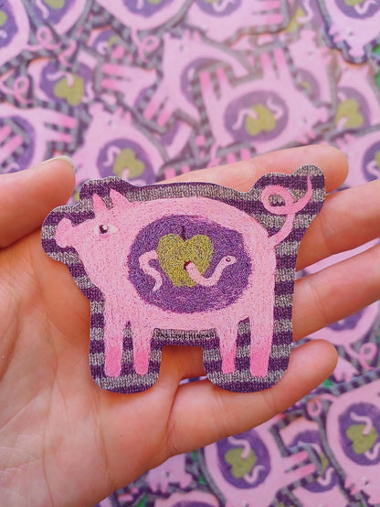 A hand holding a pink and purple felt pig-shaped sticker with a green heart in the center, against a background of multiple matching felt pig cutouts.