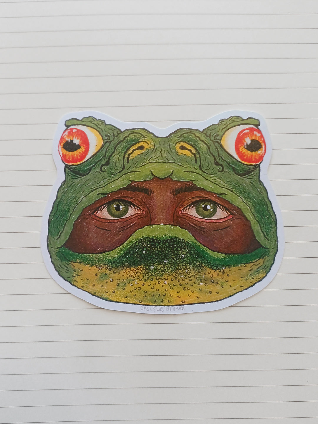 A die-cut sticker featuring a surreal frog head illustration with human eyes and a human face inside the frog's mouth, placed on a white lined notebook page.