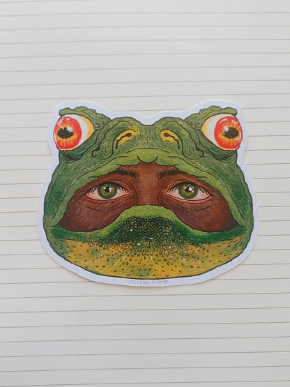 A die-cut sticker featuring a surreal frog head illustration with human eyes and a human face inside the frog's mouth, placed on a white lined notebook page.