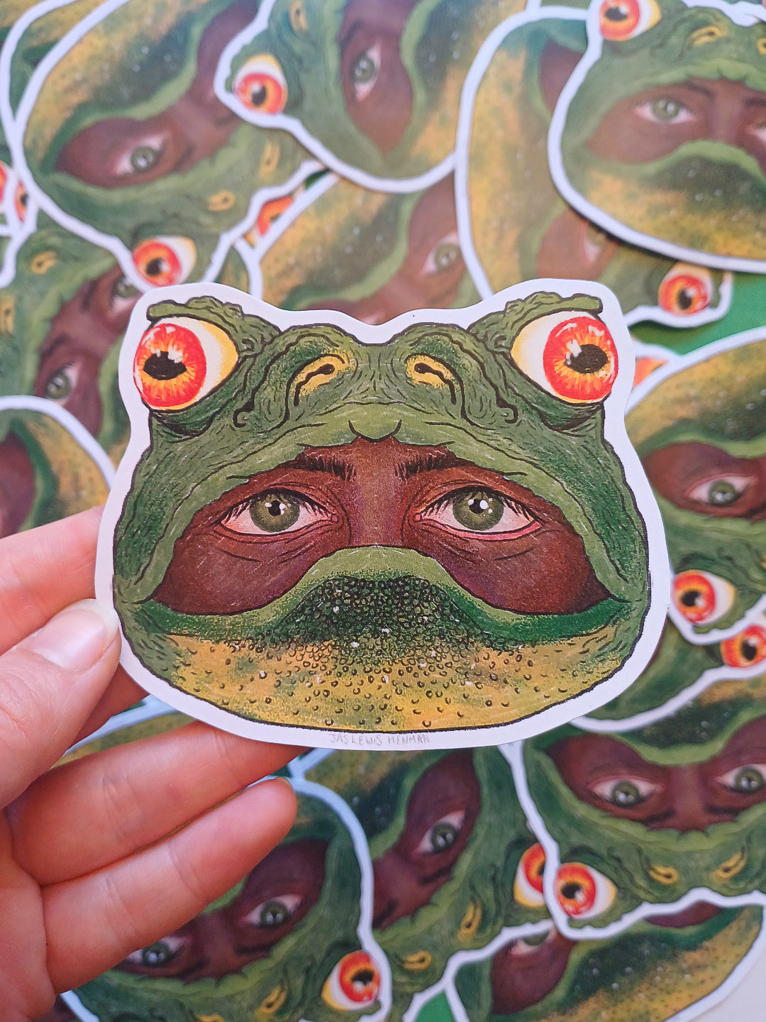 A hand holds a die-cut sticker shaped like a frog head with large human eyes in its mouth, with multiple matching stickers scattered in the background.