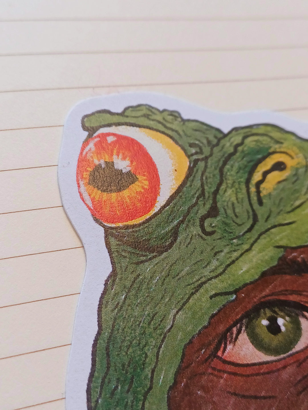 A close-up of a die-cut sticker featuring a surreal green toad with one large orange-red eye and part of another green eye, placed on a beige lined notebook background.
