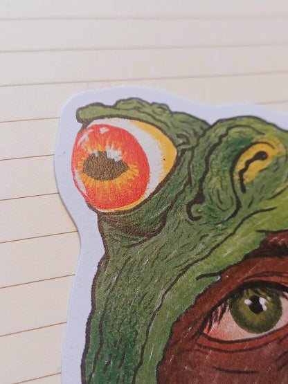 A close-up of a die-cut sticker featuring a surreal green toad with one large orange-red eye and part of another green eye, placed on a beige lined notebook background.