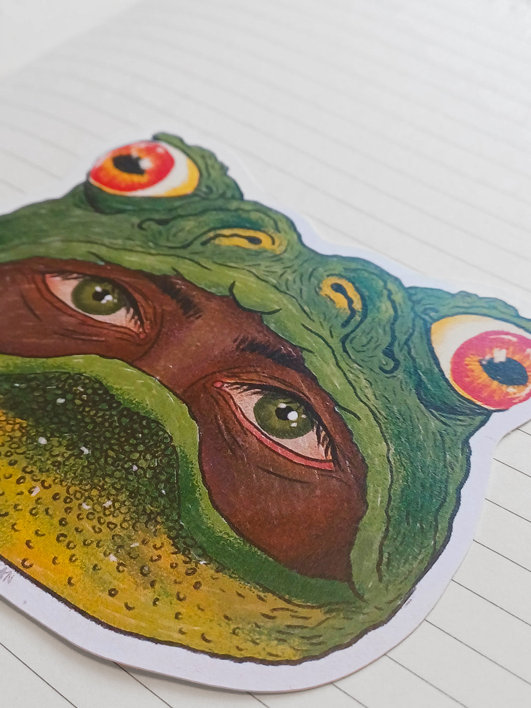 A close-up of a die-cut sticker featuring a surreal frog face illustration, placed on a white lined notebook page.
